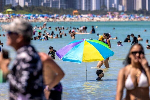 Un adolescent sta in adancurile oceanului sub o umbrela viu colorata infipta in nisip, cu multimi de oameni in jurul lui in apa.